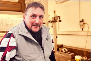 Ein Mann mit Schnurrbart und einer hellgrauen und kastanienbraunen Jacke steht in einem Labor oder einer Werkstatt mit technischen Geräten und Diagrammen im Hintergrund.