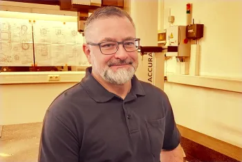Ein Mann mit Brille und grauem Bart, der ein dunkles Poloshirt trägt, steht lächelnd in einer hell erleuchteten Werkstatt oder einem Labor mit technischen Zeichnungen und Geräten im Hintergrund.