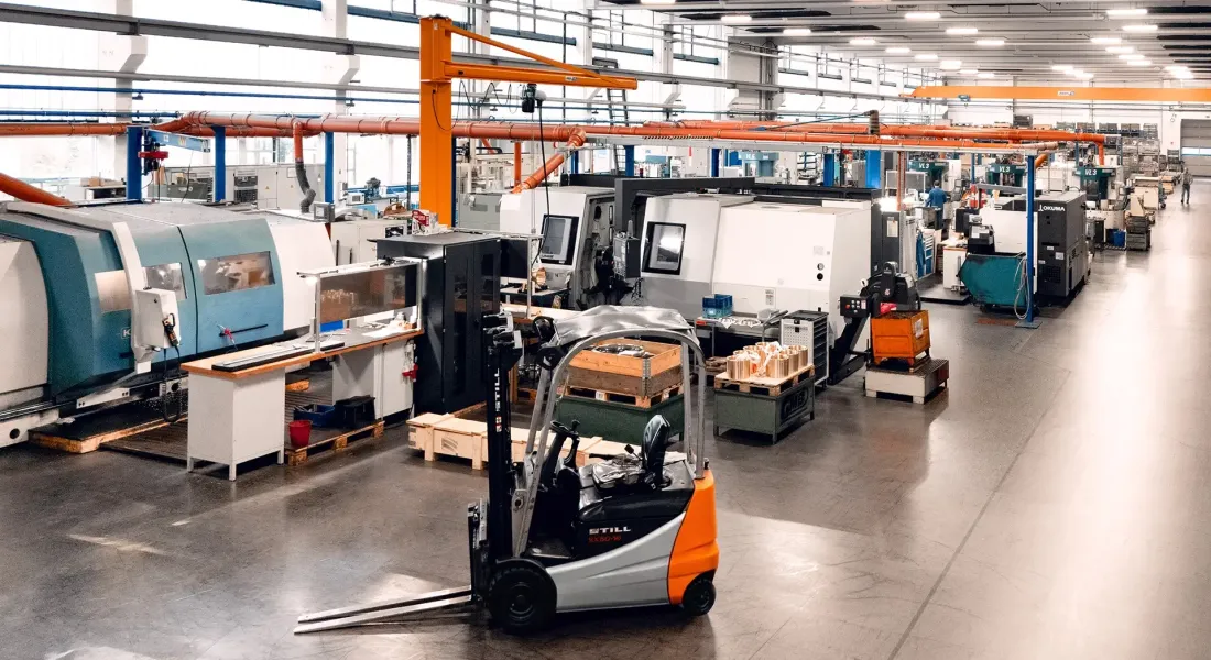 Ein Gabelstapler steht auf dem Boden einer geräumigen, gut beleuchteten Industriehalle, die mit Maschinen, Arbeitsplätzen und Geräten gefüllt ist und deren Wände mit großen Fenstern versehen sind.