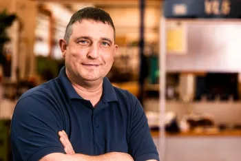 Ein Mann mit kurzen dunklen Haaren, der ein marineblaues Polohemd trägt, steht mit verschränkten Armen und lächelt in einem hell erleuchteten Innenraum, möglicherweise einer Werkstatt oder einer Industrieumgebung.